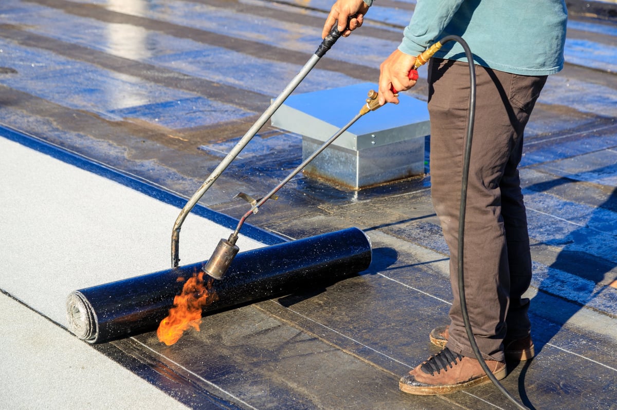 A roofer torches a membrane layer on a flat roof using a tiger torch to seal the asphalt bitumen